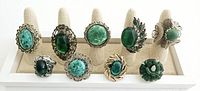 Nine vintage/antique style cocktail rings with various green stones and silver-tone settings on cream display stand