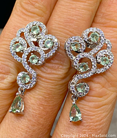 Close-up of front side of earrings worn on fingers showing the swirling scroll design, clear stones, and green gemstones including pear-shaped drops.