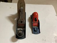 Photo shows two Stanley hand planes side by side on a white surface. The larger No. 4 plane has natural wood handles and a metal body; the smaller No. 9 block plane has a red top and black metal body.