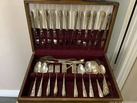 Full view of the silver plated flatware set arranged in the wooden box with velvet interior showing knives in the lid and forks, spoons and other utensils in the bottom.