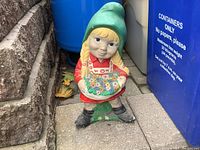 Front view of concrete little girl garden statue showing painted details including red dress, green hat, blonde braids, and colorful flower basket.