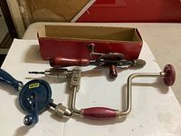 Three vintage hand drills laid out on a surface, showing wooden and metal handles, gears, and crank handles.