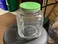 Full view of the glass cookie jar with green metal lid placed on a round stool, showing the shape, material, and lid condition.