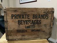 View of one side of the wooden beverage box showing black stenciled text reading 'PRIVATE BRANDS BEVERAGES LIMITED TORONTO ONTARIO' on natural wood surface.