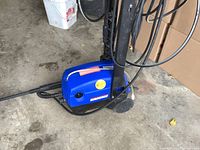 Blue Simonize PowerWash electric pressure washer positioned on a concrete garage floor, showing the side with brand name visible, attached hose wrapped around the handle, and long spray wand lying outstretched.