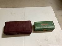 Closed maroon hard case labeled Singer on top, and green Singer cardboard box, both on white background.