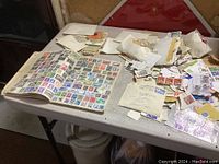 Wide shot of the full stamp album opened next to piles of envelopes and loose stamps on a table.