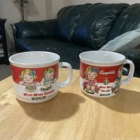 Two vintage Campbell Soup mugs/bowls with children cartoon designs placed on a wooden table in front of a dark green couch.