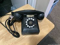 Front angle photo of black rotary dial phone with handset and coiled cord on wooden surface.