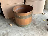 Side view of vintage copper pail with visible brass bands and a metal handle sitting on a concrete floor near cardboard boxes.