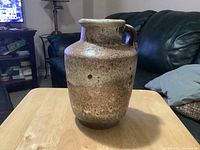 Front-facing view of a vintage stoneware jug with a single handle and wide mouth on a wooden table in a living room.
