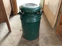Full view of the green vintage milk can standing on a concrete floor, showing its cylindrical shape and top lid with handles.
