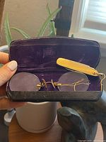 Top view of open black leatherette case lined in purple velvet containing gold-tone folding pince-nez eyeglasses and folding cutter