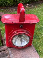 Front angled view of the red miner's lantern showing front circular glass lens and metal body with top handle.