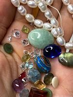 Hand holding various loose gemstones, a pearl strand, and a blue topaz silver pendant.