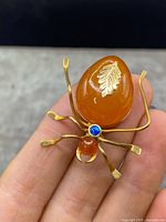 Front view of entire spider brooch held between fingers, showing teardrop amber abdomen with leaf detail, small round amber head, opal accent and bent legs.