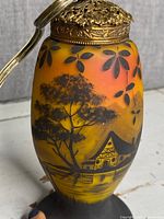 Close-up of glass lamp body showing detailed reverse painted landscape with trees and house, and ornate metal top