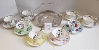Photo showing eight assorted china teacups and saucers in various floral designs and colors arranged around a silverplate basket.