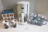 Krups white 10-cup coffee maker with glass carafe and six coffee mugs: two white, two blue from Crate & Barrel, and two cat-themed Roy Kirkham mugs
