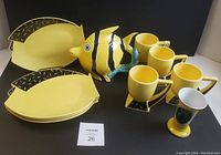 Set of 4 yellow fish-shaped plates, 4 matching mugs, fish-shaped teapot, and yellow vase on black background.