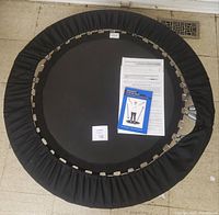 Top view of the circular black Needak Softbounce rebounder with the book and paper placed on the jumping surface, showing metal springs and black protective cover.