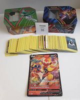 Two Pokémon card tins: a green tin with Gallade and a blue tin with Pokémon Sword and Shield artwork, with stacks of cards spread out in front including a large card.