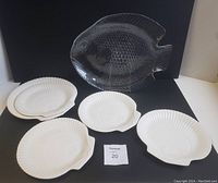 Full lot showing one large glass fish-shaped platter behind one large white Wedgwood shell plate and four smaller white Wedgwood shell plates in front.