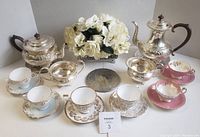 Full lot showing silver plate tea service with coffee pot, teapot, cream and sugar bowls, china teacups and saucers, trivets, and faux floral centerpiece