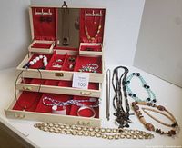 Overview of jewelry box opened showing multiple compartments with costume jewelry items including rings, earrings, necklaces, and bracelets, plus several necklaces arranged outside the box.