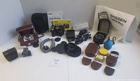 Overview of all lot items with Nikon CoolPix 5000 camera, Balda Prontor SVS camera, Goshen Luna-Pro light meter, manuals, memory cards, lens filters, and other camera accessories laid out on white surface