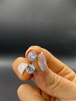 Front view of two large round-cut cubic zirconia stud earrings held between fingers, showing the stones and gold setting.