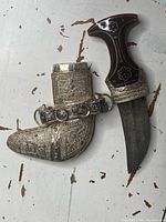 Antique Middle Eastern dagger alongside silver sheath with intricate designs on a worn surface.