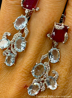 Close-up photo showing pendant earrings with red rectangular rubies and multiple oval blue topaz gemstones set in sterling silver with diamond accents.