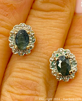 Close-up of earrings on fingers showing oval emerald stones and detailed silver and gold gilt overlay setting.