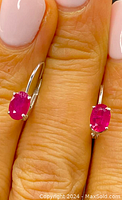 Close-up view of pair of earrings held between fingers, showing vibrant oval rubies set in 14k white gold prong settings with lever-back hooks.