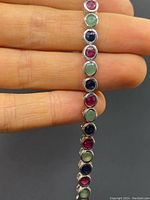 Close-up photo of bracelet held between fingers showing round rubies, sapphires and emeralds set in sterling silver.