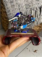 Front angled view of silver and enamel Chinese ornament showing two birds on branches with flowers on a reddish curved wooden stand held in hand.