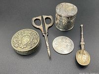 Overall view of antique silver items including scissors, silver coin, ornamental spoon, oval box, and cylindrical container