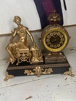 Full view showing gold-tone seated woman figure next to ornate clock face on dark base with gold decorative feet and accents.