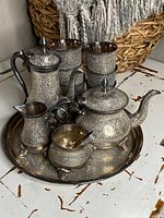 Photo showing full tea service set including tray, teapot, coffee pot, sugar bowl with spoon, creamer, and four cups, all with intricate carved floral patterns.