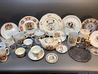 Group view of all ceramic and metal commemorative items with Queen Elizabeth II images including plates, mugs, teapot, and decorative items.