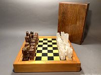 Chessboard with carved brown and white stone chess pieces set up for play. A dark stained wooden box is behind the set.