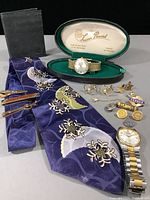 Overview of lot showing watches, tie, wallet, cufflinks, tie clips, buttons and pin arranged on gray surface