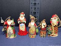 Front view of 5 Jim Shore Heartwood Creek Santa ornaments ranging from about 4 to 5 inches tall with distinct holiday designs and twine hanging loops