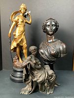 Photo showing three statuaries together: standing golden archer, dark plaster bust on wood base, seated woman statue.
