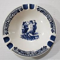 Top view of oval white ceramic ashtray decorated with Delft Blue blue patterns and a central scene with two men smoking by a stone monument, showing four cigarette rests painted blue and a decorative floral border.