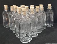 16 clear glass apothecary-style bottles with natural cork stoppers arranged together on black background.