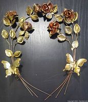 View of both floral sculptures side by side showing copper roses, brass leaves, and butterflies, laying on black surface.