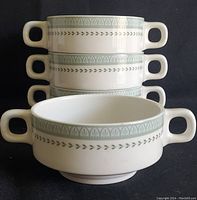 Four stacked white porcelain double handled soup bowls with a decorative light green band and leaf motif.