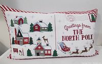 Front view of the rectangular holiday cushion showing embroidered North Pole greetings, Santa sleigh, village houses, trees, and reindeer on white fabric with red and white striped piping edges.